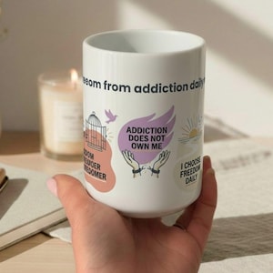 Peut inclure: Mug en céramique blanche avec l'inscription "freedom from addiction daily". La tasse présente des illustrations d'une cage à oiseaux, de mains et d'un lever de soleil, ainsi que les phrases "Addiction does not own me" et "I choose freedom daily."