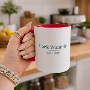 Op de afbeelding: Een witte keramische mok met een rode binnenkant en handvat. Op de voorzijde van de mok staat de donkergroene tekst Cock Womble boven een decoratieve lijn en de tekst Est. Daily. De mok heeft een gladde afwerking en een cilindrische vorm.