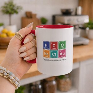 Peut inclure: Un mug en céramique blanc avec un intérieur et une anse rouges, orné d'un graphique de tableau périodique avec les éléments H, C, O, Na, Cl et Au. Sous les carrés colorés, le texte Your Custom Name Here est écrit en police noire.