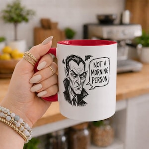 Peut inclure: Un mug en céramique blanche avec un intérieur et une anse rouges, présentant une illustration graphique noire d'un vampire et une bulle contenant le texte NOT A MORNING PERSON. Le mug a une finition lisse et une forme cylindrique.