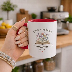 Peut inclure: Un mug en céramique blanche avec un intérieur et une anse rouges. L'avant présente une illustration d'un livre ouvert avec des fleurs sauvages colorées sortant des pages. Le texte indique : A fresh story blooms, mama, est. 2024, avec un petit cœur rouge.