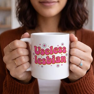 May include: White ceramic mug with the text "Useless lesbian" in pink and orange retro font, decorated with small daisy and star graphics. The mug is held by a person.