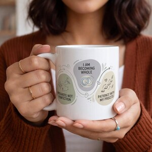Könnte beinhalten: Weiße Keramik-Tasse mit den Aufschriften "I Am Becoming Whole", "Progress Not Perfection" und "Patience With My Process". Die Tasse hat einen Henkel und ist mit einem hellgrünen, grauen und beigen Design verziert.