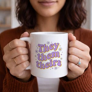 Puede incluir: Taza de cerámica blanca con las palabras "They them theirs" en una fuente retro morada y amarilla, decorada con pequeñas margaritas y estrellas. La taza es sostenida por una persona.