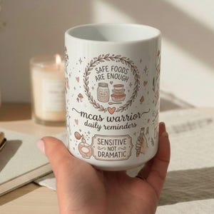 以下が含まれることがあります： イラストとテキストが入った白いセラミックマグカップ。「SAFE FOODS ARE ENOUGH」、「mcas warrior daily reminders」、「SENSITIVE NOT DRAMATIC」の文字がデザインされています。食べ物、ハート、その他の装飾要素の図が含まれています。