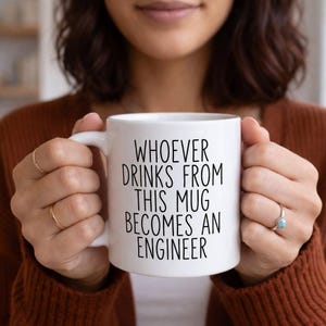 Peut inclure: Mug en céramique blanche avec l'inscription noire "WHOEVER DRINKS FROM THIS MUG BECOMES AN ENGINEER". La tasse est tenue par une personne portant un pull marron. La tasse a une anse blanche.