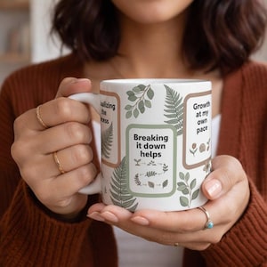Peut inclure: Mug en céramique blanche avec des illustrations botaniques et les phrases « Breaking it down helps » et « Growth at my own pace ». La tasse a une anse et est décorée d'accents verts et bruns.