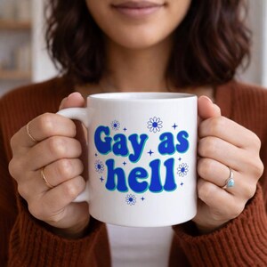 Può includere: Tazza in ceramica bianca con la scritta "Gay as hell" in blu e azzurro, decorata con grafiche floreali e stellate. La tazza ha un manico bianco ed è tenuta da una persona.