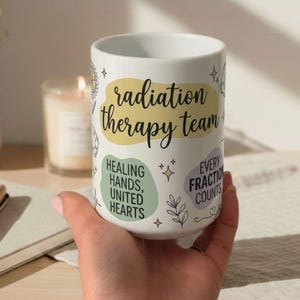 Peut inclure: Mug en céramique blanche avec l'inscription "radiation therapy team" dans une police manuscrite. La tasse présente également les phrases "Healing Hands, United Hearts" et "Every Fraction Counts" dans un design fantaisiste.