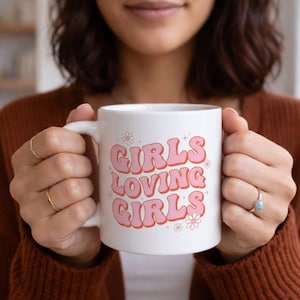 Puede incluir: Taza de cerámica blanca con el texto "GIRLS LOVING GIRLS" en letras rosas con burbujas. La taza tiene un asa blanca y está decorada con pequeños gráficos de flores. La taza está sostenida por una persona.