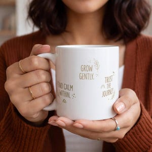 Peut inclure: Mug en céramique blanche avec les phrases « Grow Gently », « Find Calm Within » et « Trust the Journey ». La tasse a une anse simple et est tenue par une personne. Le design est minimaliste.