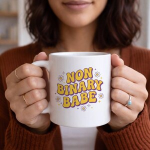 Puede incluir: Taza de cerámica blanca con el texto "NON BINARY BABE" en amarillo y morado, decorada con pequeñas ilustraciones de margaritas. La taza tiene un asa curva y está sostenida por una persona.