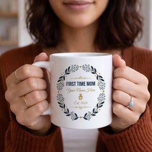Peut inclure: Mug en céramique blanche avec une anse, orné de l'inscription "FIRST TIME MOM" et "Your Name Here" dans une couronne florale bleu marine et dorée. La tasse comprend également le texte "Est. 2026" et une icône de bébé.