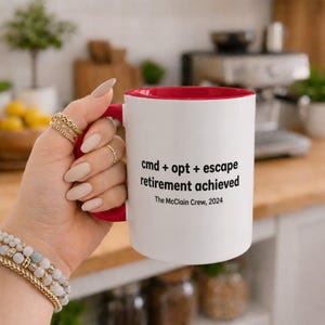 Peut inclure: Une tasse en céramique blanche avec un intérieur et une anse rouges. L'extérieur présente un texte noir indiquant : cmd + opt + escape retirement achieved, suivi de The McClain Crew, 2024. La tasse est tenue par une main ornée de bagues et bracelets.