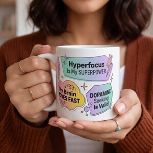 Peut inclure: Mug en céramique blanche avec les phrases "Hyperfocus Is My SUPERPOWER", "My Brain MOVES FAST" et "DOPAMINE Seeking Is Valid" en texte coloré. La tasse a une anse incurvée et est tenue par une personne.