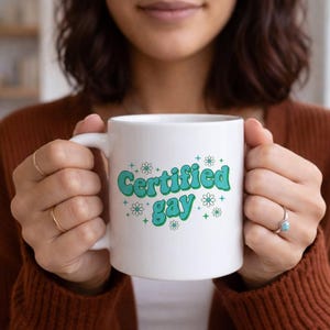 May include: White ceramic mug with the text "Certified Gay" in teal and green, surrounded by small flower and star graphics. The mug has a curved handle and is held by a person wearing a brown sweater.