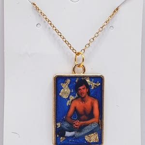 Könnte beinhalten: Goldfarbene Halskette mit rechteckigem Anhänger. Der Anhänger zeigt einen Mann in Jeans vor blauem Hintergrund mit Goldblatt-Akzenten. Der Anhänger ist goldgerahmt und hängt an einer feinen Kette. Auf der Karte steht "Handmade with love."