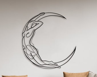 May include: A black metal wall sculpture of a woman reclining on a crescent moon. The sculpture is hanging on a white wall above a light gray sofa with brown throw pillows. The sofa is in a living room with a wooden floor and a wooden bookshelf on either side.