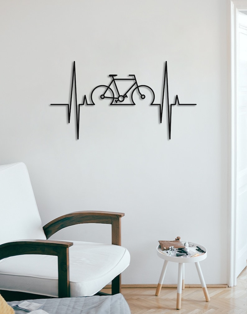 May include: Black metal wall art depicting a bicycle inside a stylized heartbeat. The art is mounted on a white wall with a white chair and a small white table with a brown top in the foreground.