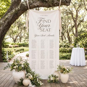 Peut inclure: Une bannière de plan de table en tissu blanc cassé suspendue à une branche d'arbre. La bannière affiche le texte V XXIX MMXIV, Find Your Seat, et Your Seat Awaits, suivi d'une liste de noms et de numéros de table. Elle est entourée de compositions florales dans des vases en céramique.