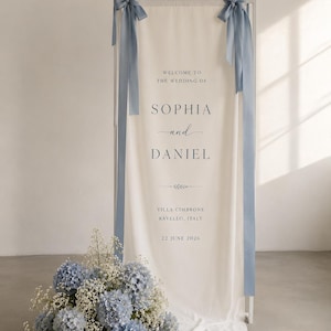 Op de afbeelding: Een hoog wit rechthoekig frame met een crèmekleurig stoffen bord met de tekst "WELCOME TO THE WEDDING OF SOPHIA and DANIEL VILLA CIMBRONE RAVELLO, ITALY 22 JUNE 2026". Twee stoffige blauwe linten zijn aan de bovenhoeken gebonden en blauwe hortensia's staan aan de voet.