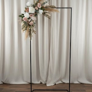 Puede incluir: Arco de boda de metal negro decorado con un arreglo floral. El arreglo presenta rosas blancas y rosas, hierba de la pampa y vegetación. El arco rectangular se apoya en un suelo de madera sobre un fondo neutro.