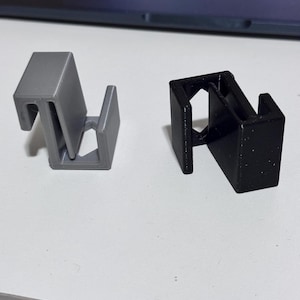 Peut inclure: Deux clips en plastique, un gris et un noir, sont présentés sur une surface blanche. Les clips ont une conception unique en forme de Z avec une base rectangulaire et un sommet en forme de crochet. Le clip gris est lisse, tandis que le clip noir a un aspect texturé.