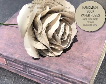 Puede incluir: Una rosa de papel hecha a mano a partir de las páginas de un libro favorito. La rosa es blanca y está sobre un libro con una cubierta morada.
