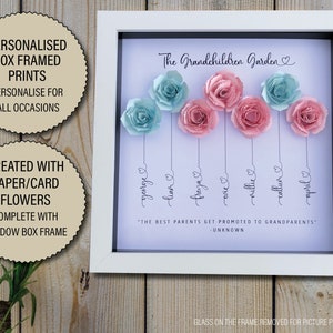 May include: A white shadow box frame with a white background and the text "The Grandchildren Garden" in black cursive. The frame contains seven paper flowers in shades of pink and blue, each with a name written below it in black cursive. The names are George, Lian, Freya, Twice, Millie, Callum, and April. The quote "The best parents get promoted to grandparents - Unknown" is printed below the flowers.