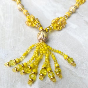 May include: A close-up of a yellow beaded necklace with a tassel pendant. The necklace features various sizes and shapes of yellow beads, with gold-coloured accents. The tassel is made of strands of yellow beads, and the pendant has a gold-coloured filigree design.