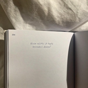 May include: An open book with the page number 254 visible. The text "Woran möchte ich heute besonders denken?" is written in black ink on the white page. The background is a soft, textured, off-white fabric.