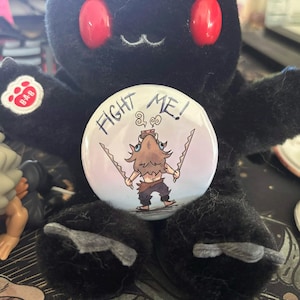 Może przedstawiać: Czarna pluszowa zabawka z czerwonymi oczami i białym guzikiem z postacią z anime i napisem "FIGHT ME!". Guzik ma kreskówkową ilustrację postaci trzymającej dwa miecze.