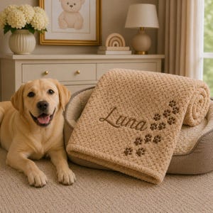 May include: A tan, textured, waffle-knit pet blanket featuring the embroidered name Luna in brown script, accompanied by a trail of brown embroidered paw prints. The blanket is folded and resting inside a tan, oval-shaped pet bed.