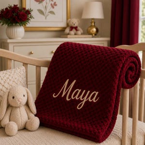 Könnte beinhalten: Eine tiefrote, waffelgestrickte Decke mit dem in beigefarbener Schreibschrift eingestickten Namen Maya. Die Decke liegt über dem Gitter eines hellen Holzbettchens neben einem kleinen, beigefarbenen Plüschhasen mit langen Ohren.