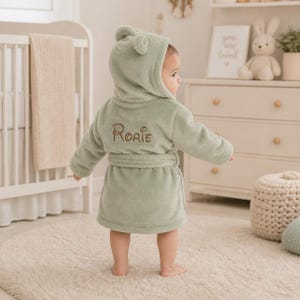 May include: A sage green plush bathrobe featuring a hood with small rounded ears and a tie belt at the waist. The back of the robe is embroidered with the name Rorie in brown thread. The fabric has a soft, fuzzy texture throughout the garment.
