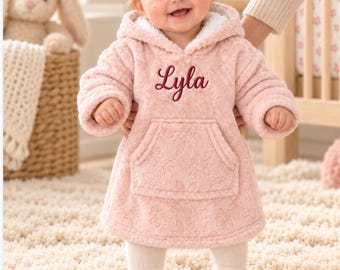 Könnte beinhalten: Ein weicher, hellrosa Kapuzenpullover aus Fleece mit weißem Sherpa-Futter, einer Kängurutasche vorne und einem geprägten Blumenmuster. Der Name Lyla ist in dunkelrosa Schreibschrift auf der Brust des Kleidungsstücks eingestickt.