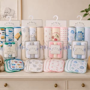 May include: A collection of Rock-a-Bye Baby Boutique bath sets featuring rolled hooded towels and square washcloths in various patterns including trucks, elephants, cherries, and blue dinosaurs, displayed with a plush bunny and a wooden sign.