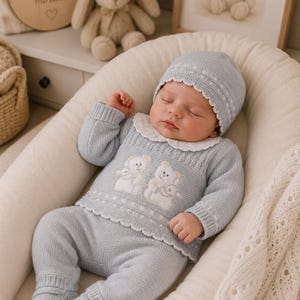 Peut inclure: Un bébé endormi portant un ensemble en tricot bleu clair avec un chapeau assorti. L'ensemble comprend des ours en peluche brodés blancs et un col et une bordure festonnés. Le bébé est dans un nid rembourré blanc.