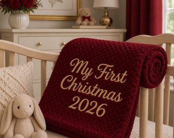 May include: A deep red, textured knit baby blanket featuring gold embroidered text that reads "My First Christmas 2026". The blanket is draped over the wooden slats of a crib, positioned next to a small, light beige plush rabbit toy.