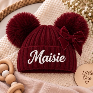 Könnte beinhalten: Eine tiefburgunderrote, gerippte Strickmütze mit zwei großen, passenden Bommeln auf der Oberseite. Die Vorderseite der Mütze ist mit dem Namen Maisie in weißer Schreibschrift bestickt. Eine dunkelrote Samtschleife ist an der rechten Bommel befestigt.