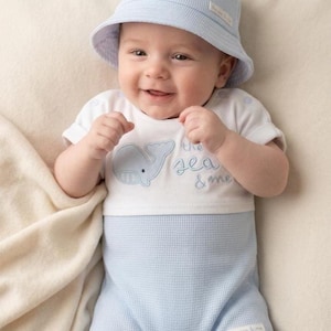 May include: A baby wearing a light blue bucket hat and a matching romper. The romper has a white top with a whale graphic and the text "the sea & me". The outfit is made of a textured fabric.