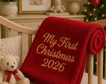May include: A deep red, textured, plush baby blanket featuring gold embroidered text that reads My First Christmas 2026. The blanket is folded and draped over a light-colored wooden crib, showcasing a soft, bumpy fabric surface.