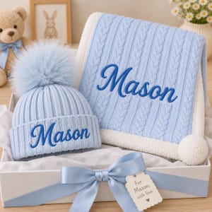 May include: A light blue cable knit baby blanket with a white sherpa lining and a matching light blue ribbed knit beanie with a large faux fur pom-pom. Both items feature the name Mason embroidered in dark blue script text.