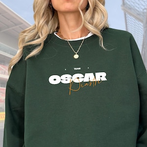 May include: Dark green sweatshirt with the text "TEAM OSCAR Piastri" in white and orange. The sweatshirt has a white collar and is worn with a gold necklace. The design is centered on the chest.