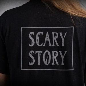 May include: Black t-shirt with a white rectangle on the back that says "SCARY STORY" in a distressed font.