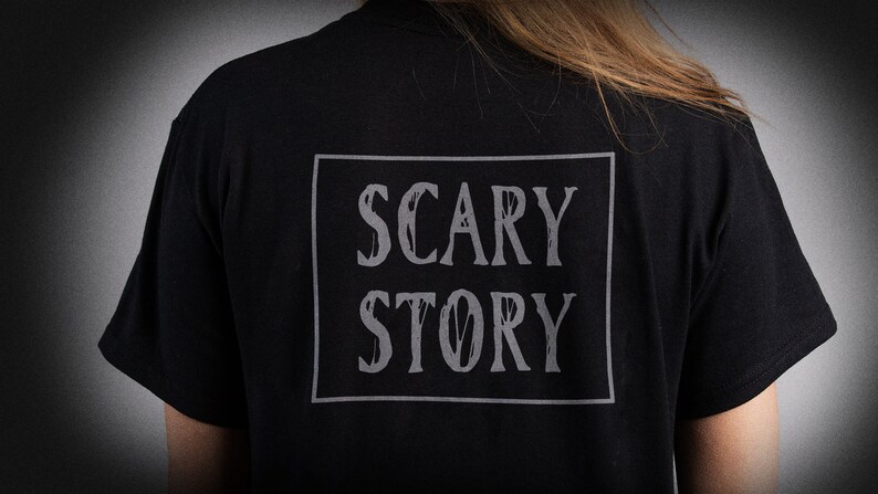 May include: Black t-shirt with a white rectangle on the back that says "SCARY STORY" in a distressed font.