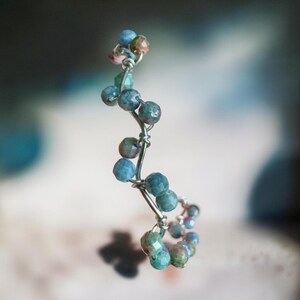 May include: A wire-wrapped ear cuff featuring a series of small, faceted, round beads in shades of blue, green, and brown. The silver-toned wire is twisted into a vertical, undulating shape that follows the curve of the beads in a continuous line.