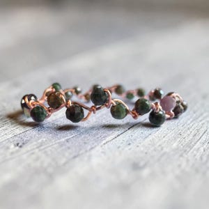 May include: A ring constructed from thin copper wire wrapped in a zigzag pattern, featuring small, faceted, dark green beads and a single light pink bead interspersed along the wire structure, creating a continuous, textured, circular band design.