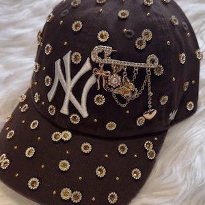 May include: A dark brown baseball cap featuring an embroidered white NY logo. The cap is decorated with numerous small, circular, gold-toned flower-shaped rhinestones and a large gold-toned safety pin brooch with dangling charms including a butterfly.