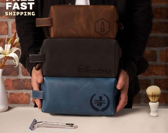 Puede incluir: Tres neceseres de cuero en marrón, negro y azul apilados. La bolsa marrón tiene las iniciales "AD" dentro de un hexágono. La bolsa negra está inscrita con el nombre "Theodore". La bolsa azul tiene la inicial "J" dentro de una corona de laurel. El texto "SAME DAY FREE FAST SHIPPING" es visible.